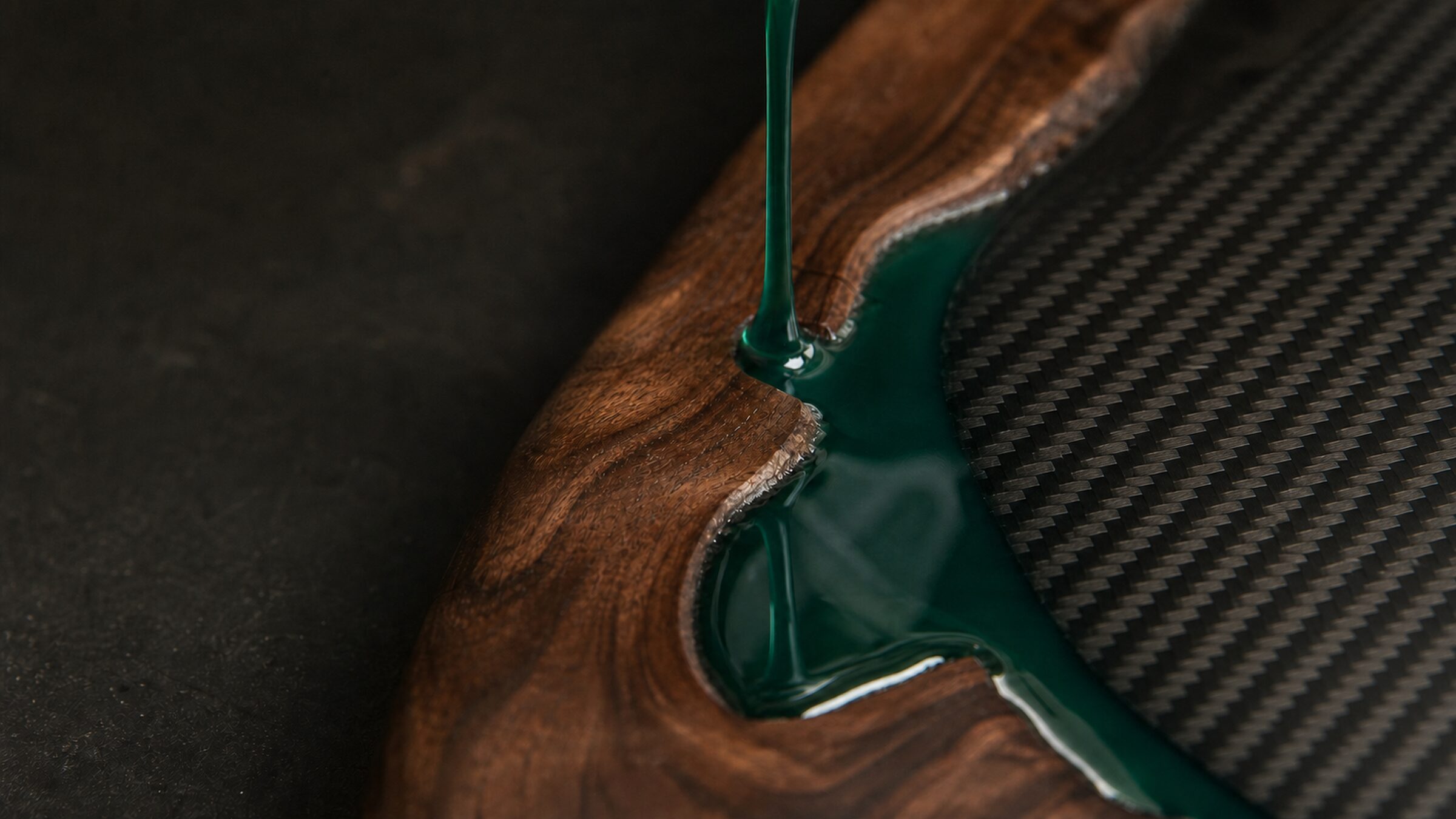 Resina epoxi verde profundo fluye sobre una trama de fibra de carbono y una veta de madera oscura.